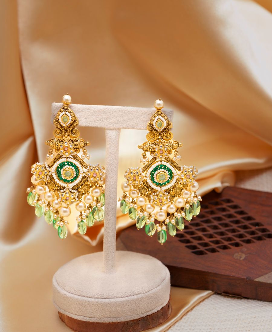 Gold chandelier earrings with green gemstone detailing against a beige background
